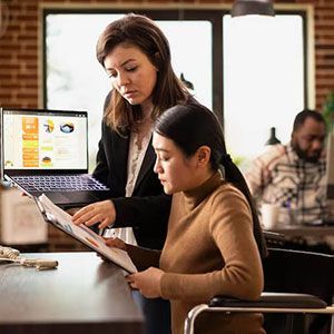Two professionals in an office collaborate, with one pointing to a document and the other holding a laptop.