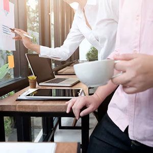 Two people collaborating in a bright office, one pointing at wall charts while the other holds a cup of coffee.