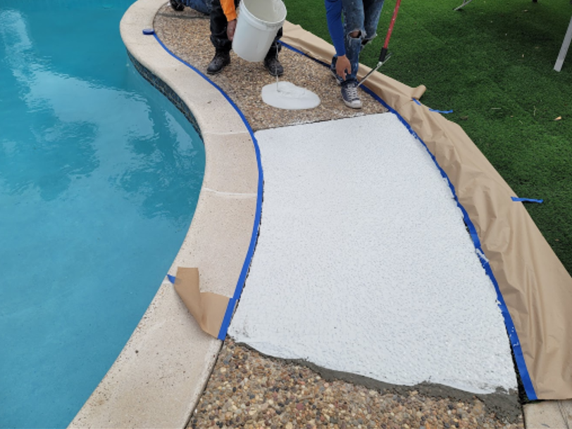 Workers pouring white coating on a pool walkway, protected by blue tape and brown paper.