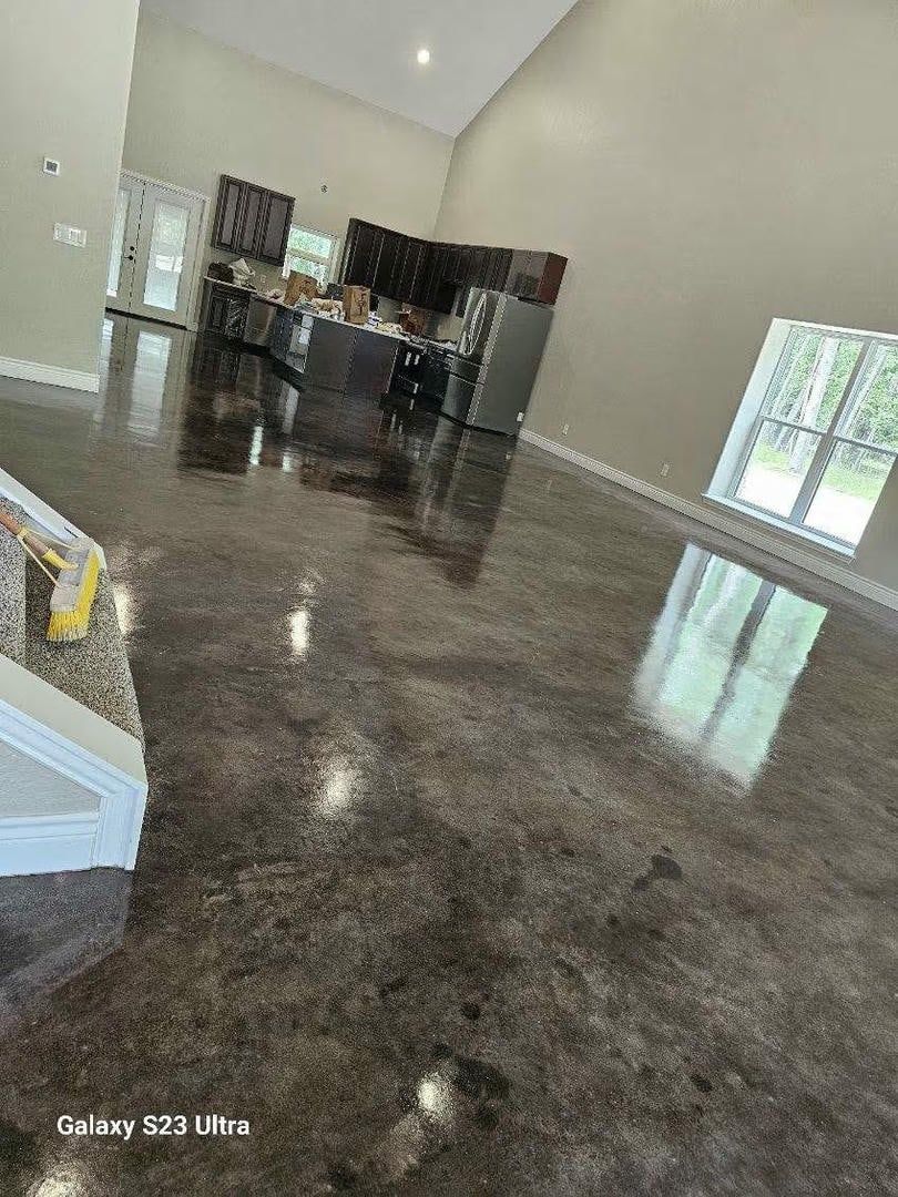 Glossy, dark-colored concrete floor in a room with tall ceilings, kitchen in background, sunlight from window.