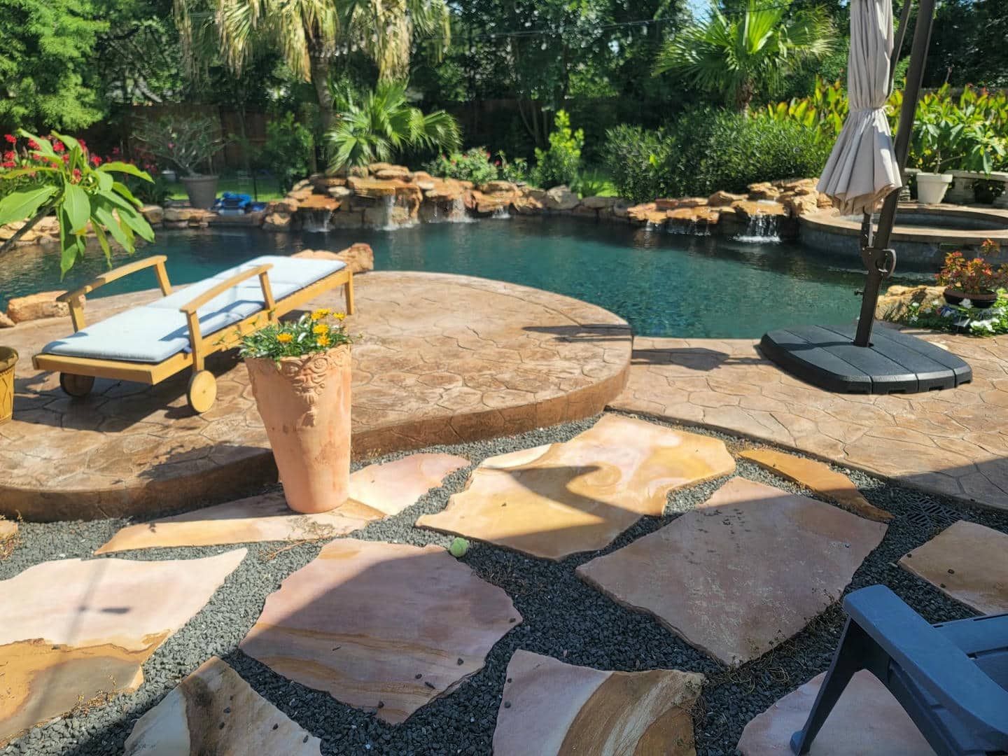 Poolside patio with irregular stone pavers, chaise lounge, and umbrella. Water features and lush greenery in the background.