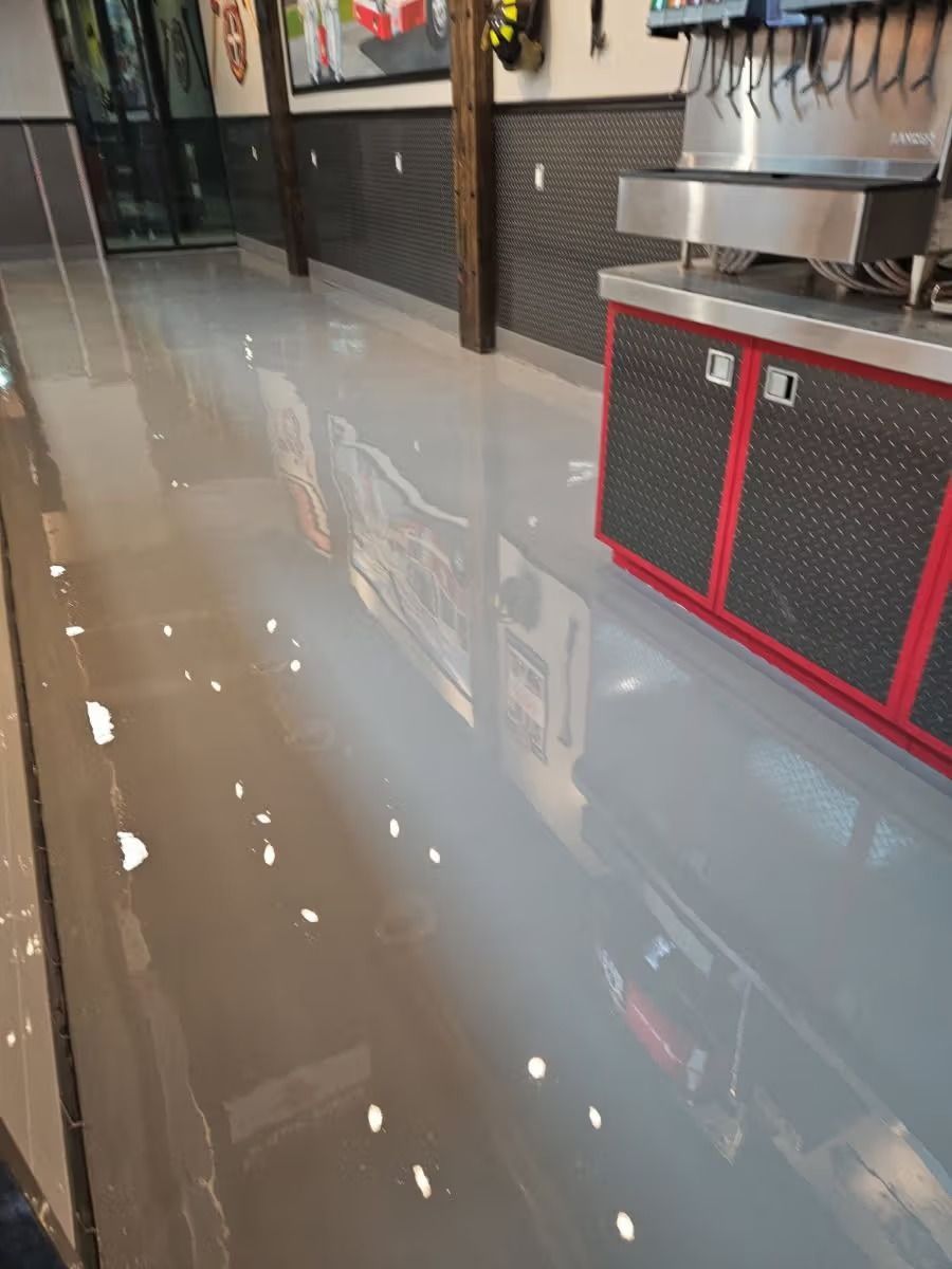Shiny, gray epoxy floor reflecting overhead lights in a commercial space near a soda machine and a wall with textured panels.