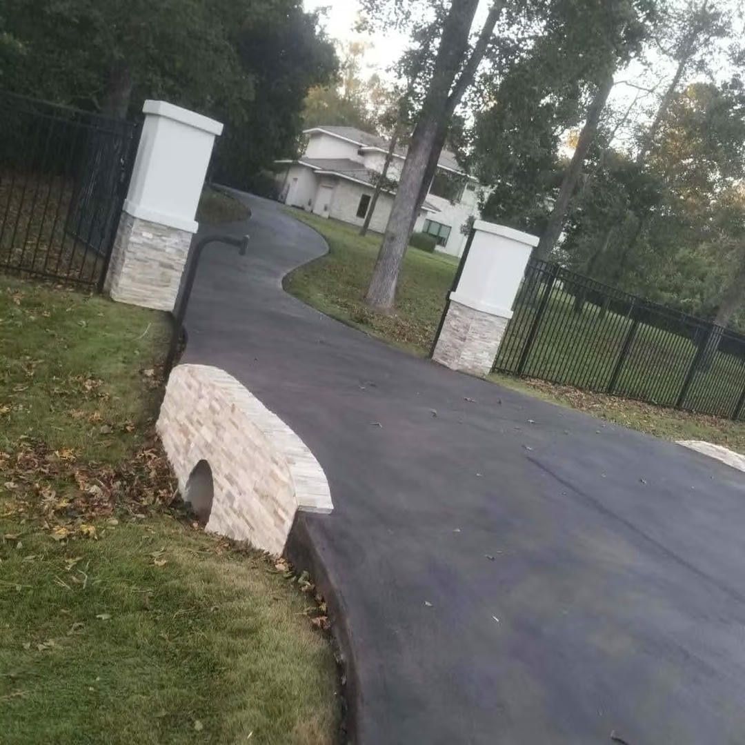 Black asphalt driveway leading to a white house, with stone pillars and black fencing on either side.