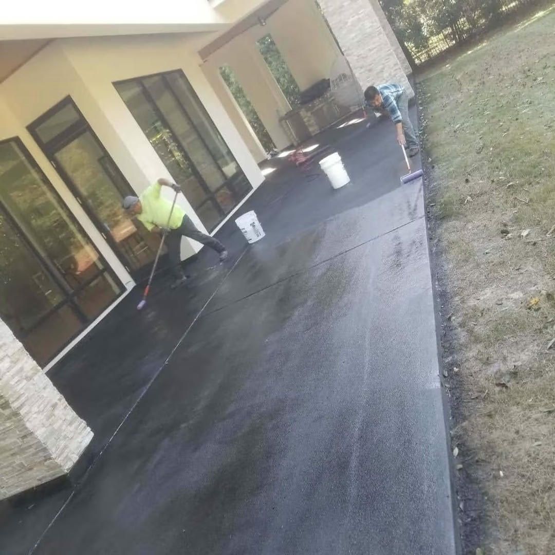 Two people applying coating to a dark gray concrete patio with rollers, beside a building with large windows.
