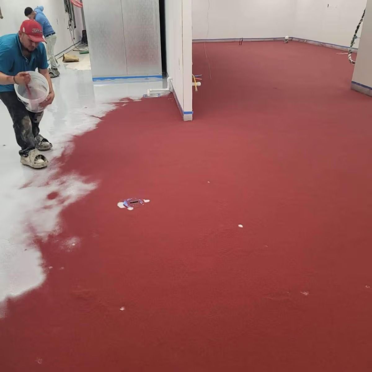 Person pouring white substance onto red floor in a room under construction.