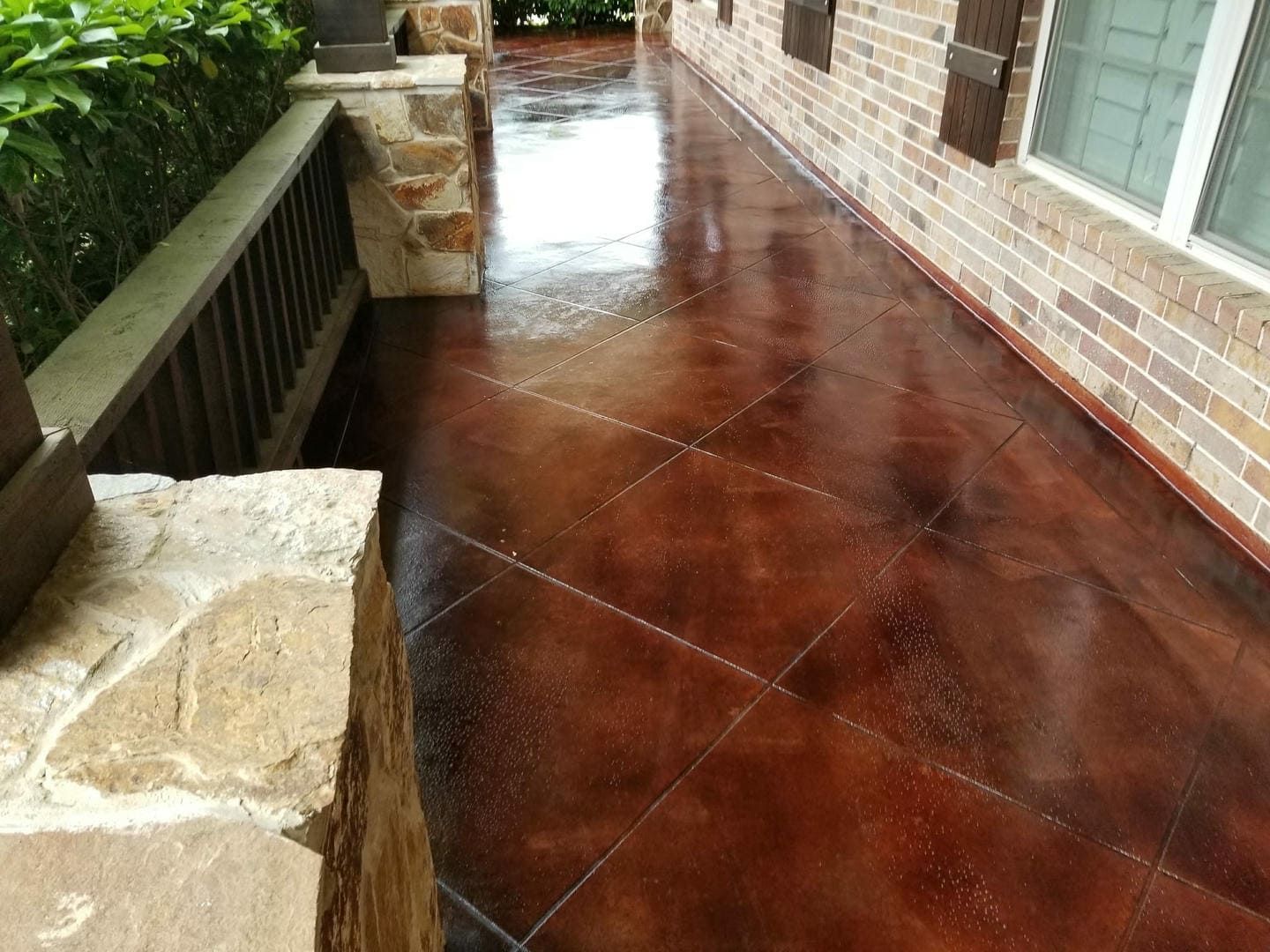 Brown stained concrete porch with tile pattern, next to brick wall and stone pillar.