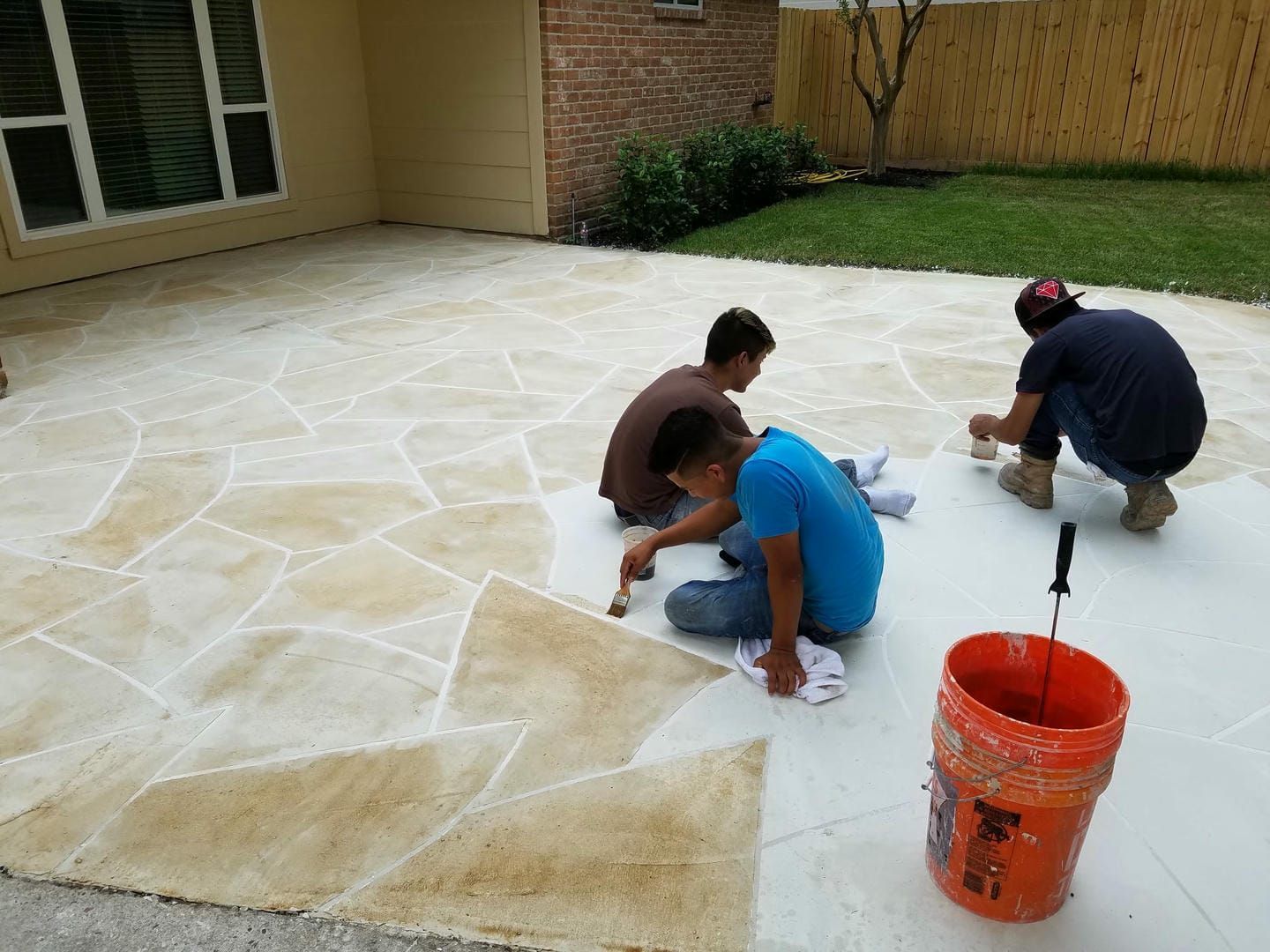 Three people painting a patio floor with a stone pattern. A bucket of paint and supplies are visible.