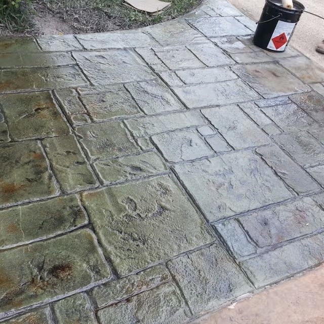 Stamped concrete patio with varying shades of gray, green, and brown, resembling flagstone. A bucket of sealant sits nearby.