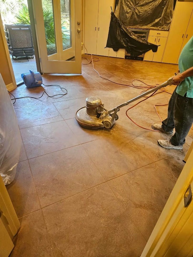 Person using a floor buffer on a brown floor; interior with open door and plastic covering.