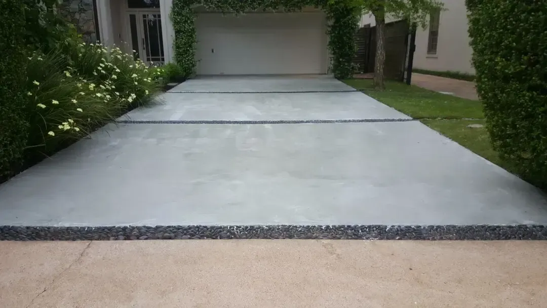 Concrete driveway leading to a garage, flanked by greenery. Black gravel border.