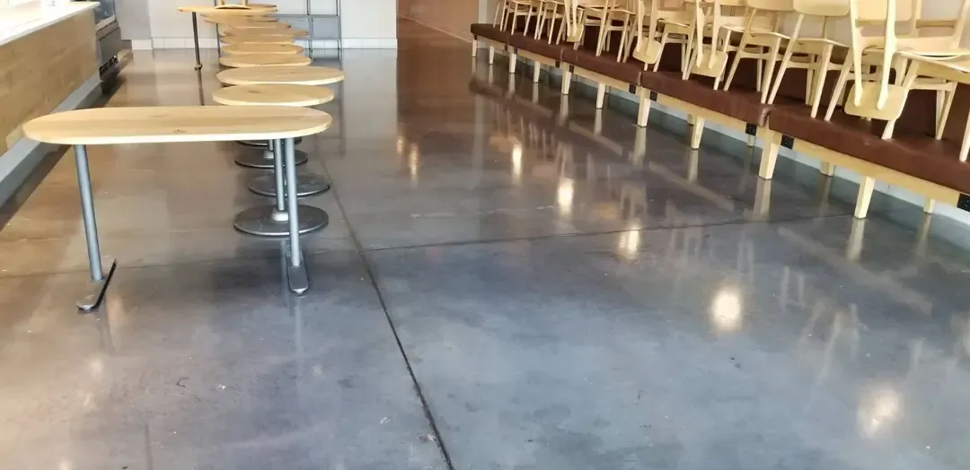 A restaurant interior featuring polished concrete floors, long tables, and rows of chairs.