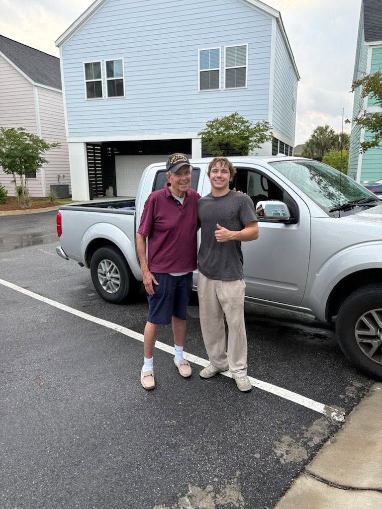 Two people standing beside a silver pickup truck in a parking lot, smiling at the camera.