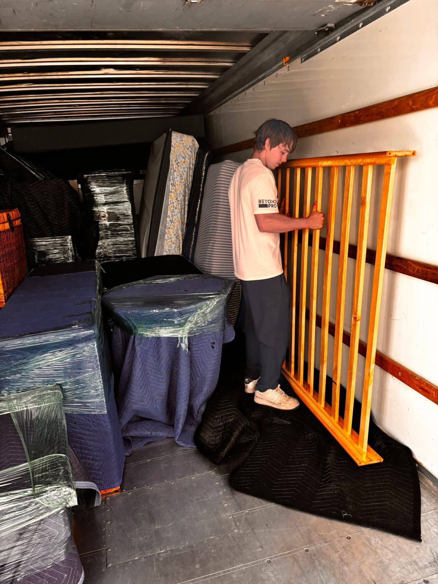 Man moving orange pallet on a truck bed beside furniture wrapped in protective blankets