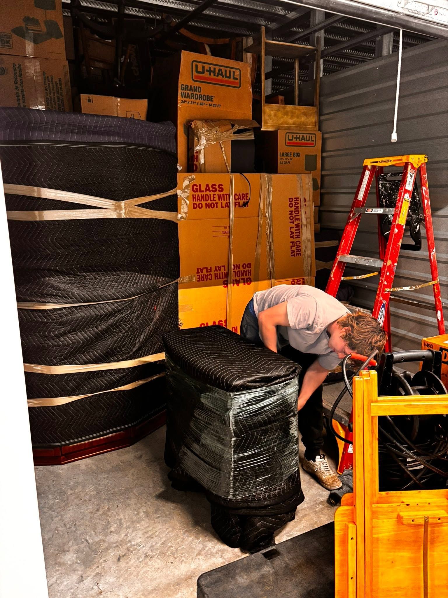 Worker bending over stacked tires in a warehouse, with boxes and a red ladder nearby.
