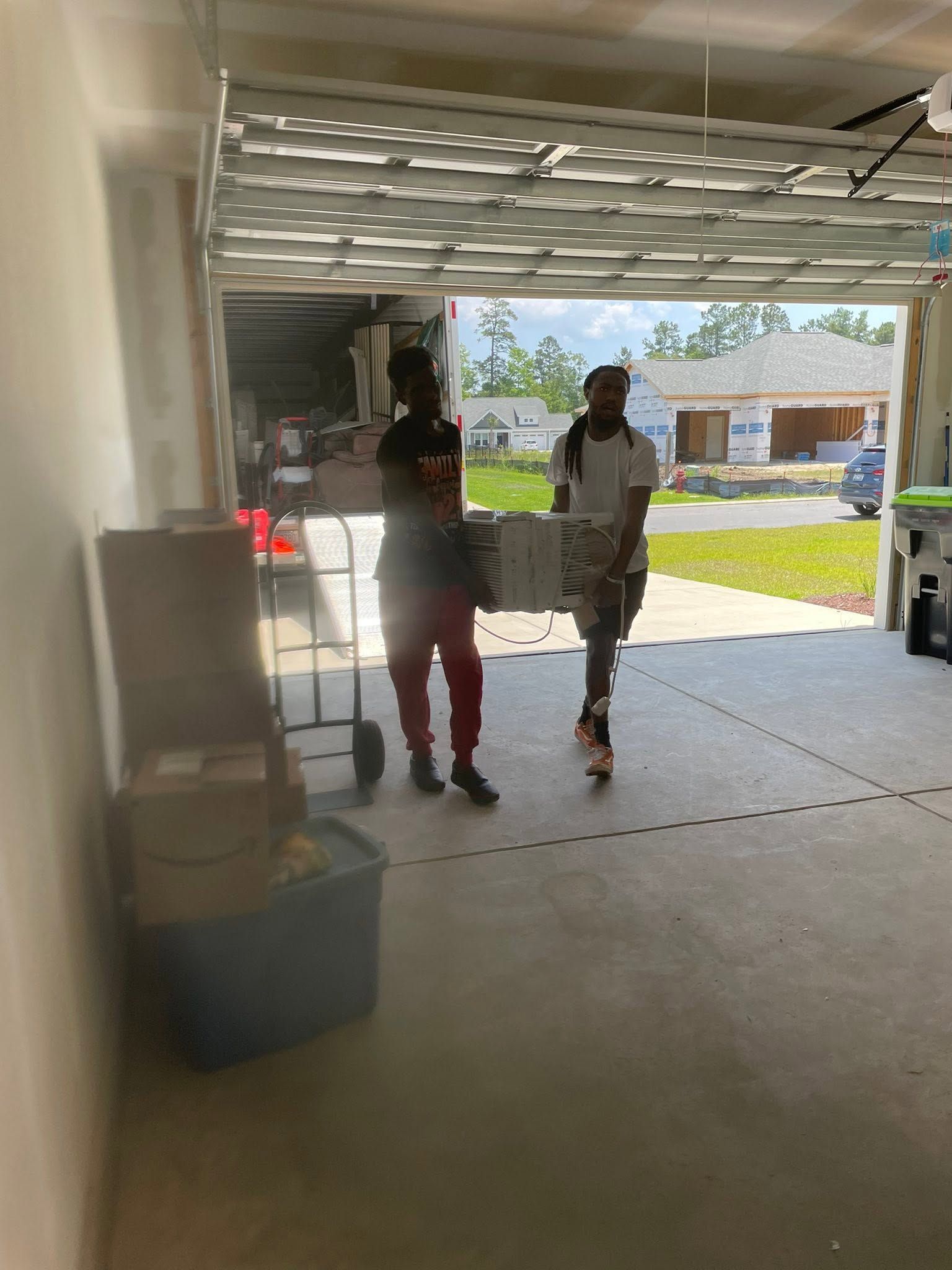 Two people standing in a garage doorway, with daylight and houses visible outside.