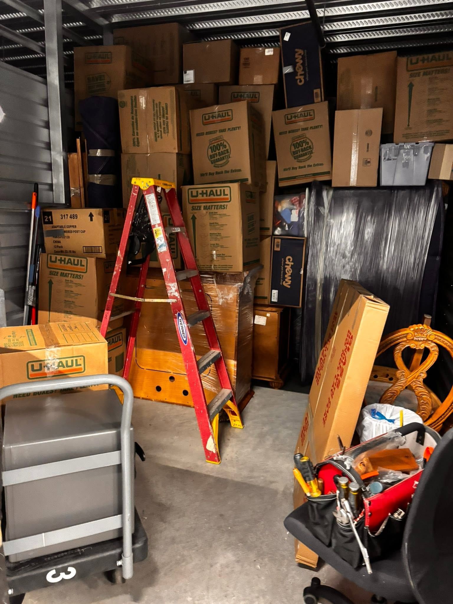 Storage room stacked with cardboard boxes, a red ladder, and assorted tools and equipment.