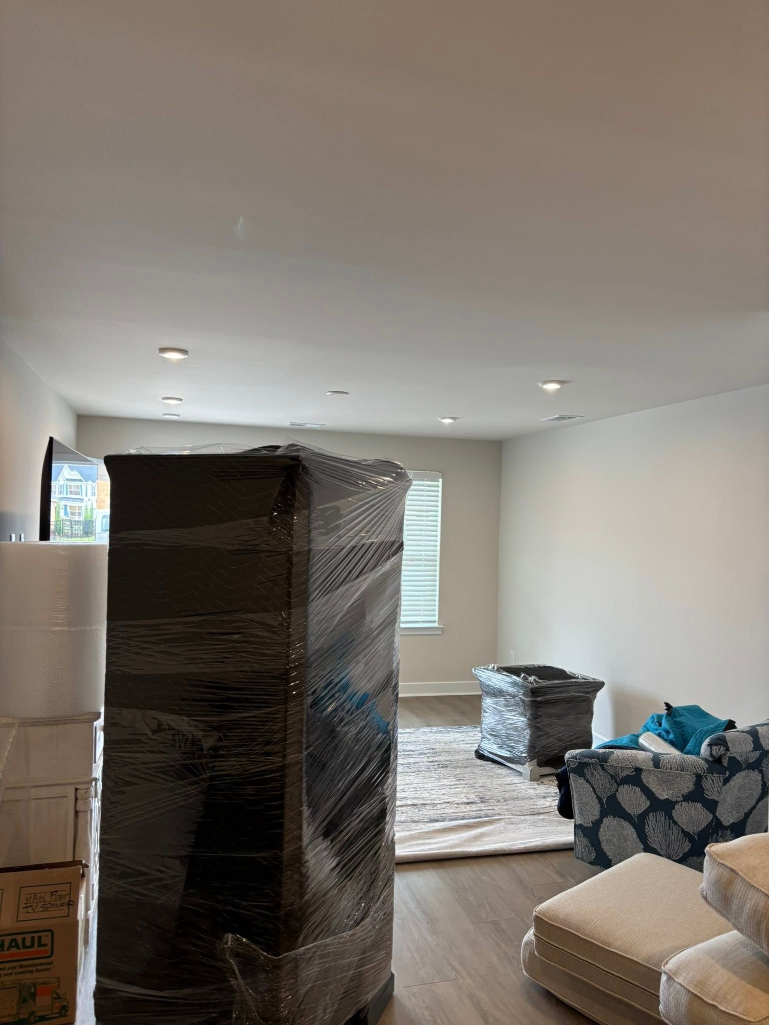Living room with stacked wrapped furniture, boxes, and moving supplies on the floor