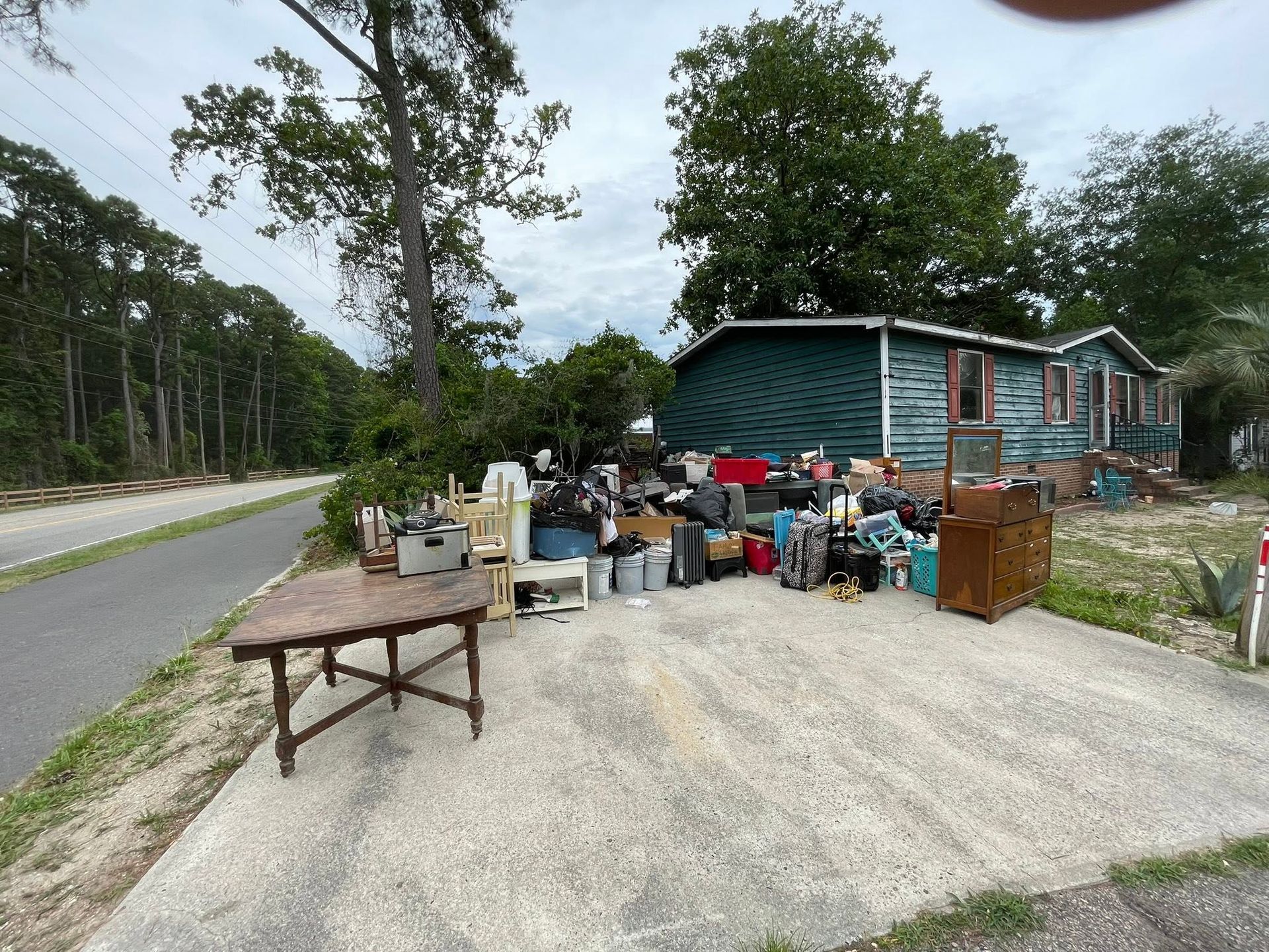 Roadside yard sale with assorted items on a gravel driveway beside a teal house and wooded road
