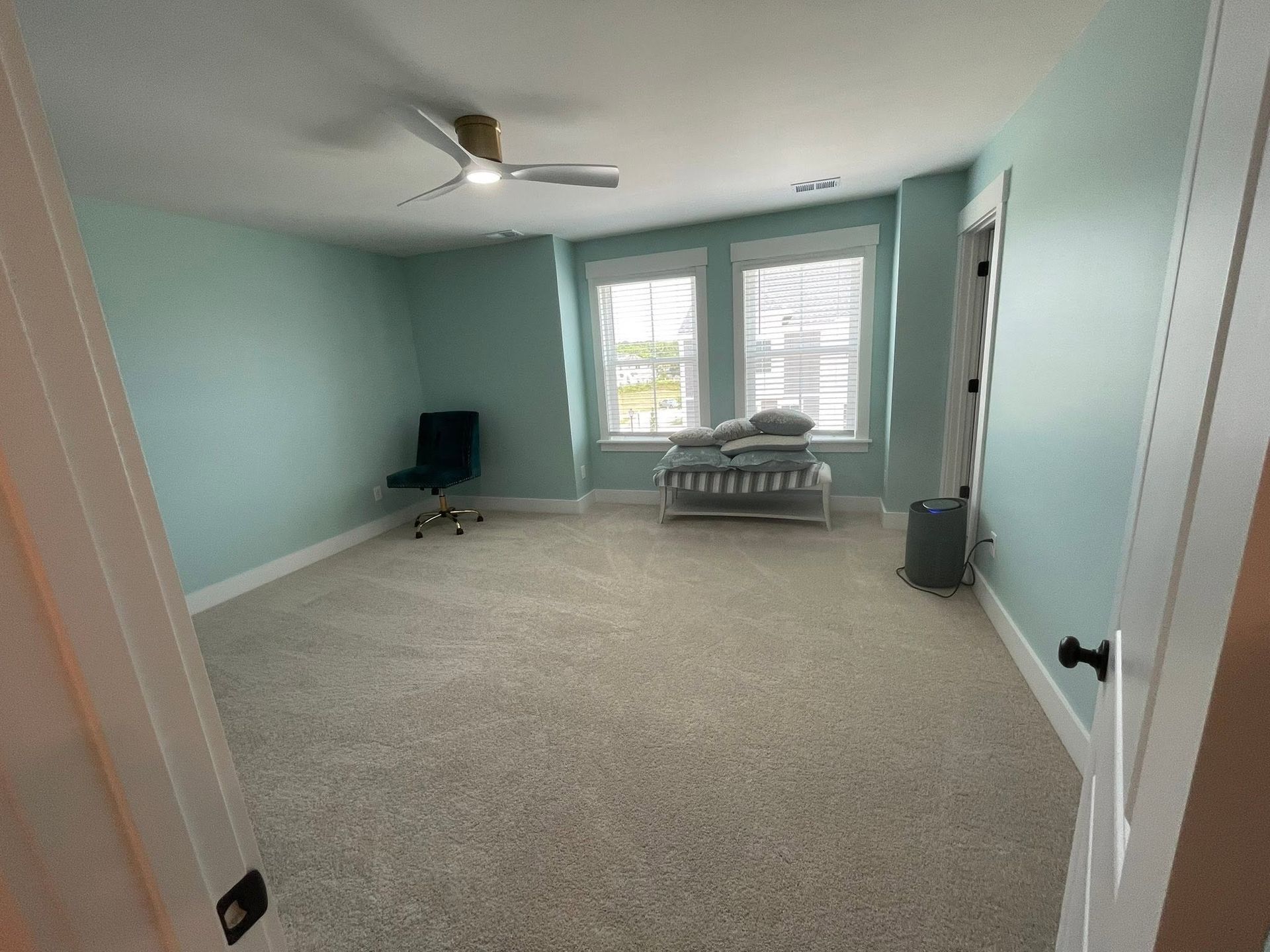 Empty light-blue bedroom with carpet, ceiling fan, window seat, and a chair near the window.