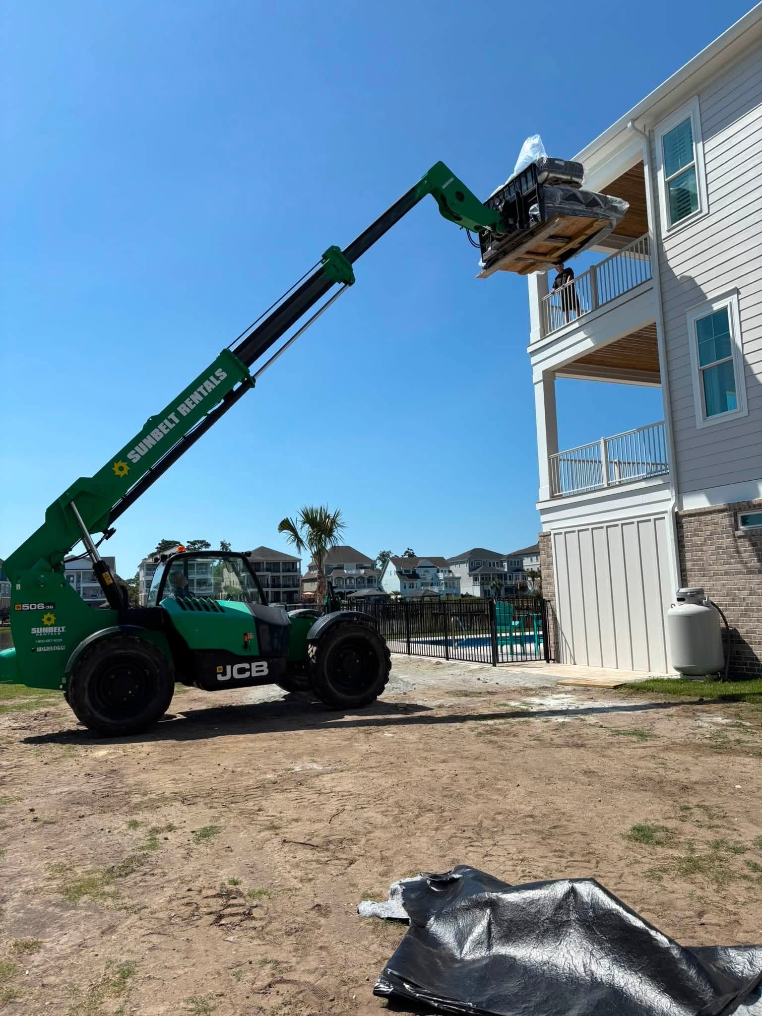 Green telehandler lifting a worker to the upper balcony of a white building on a sunny day