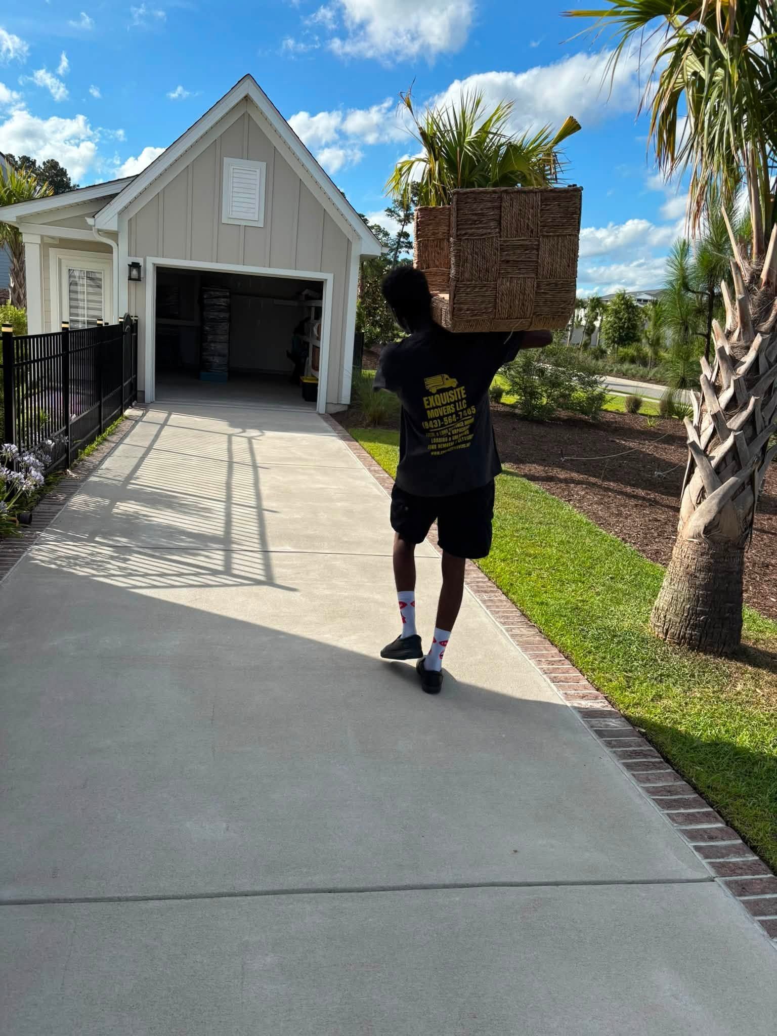 Person walking up a driveway carrying a large basket toward a house garage on a sunny day