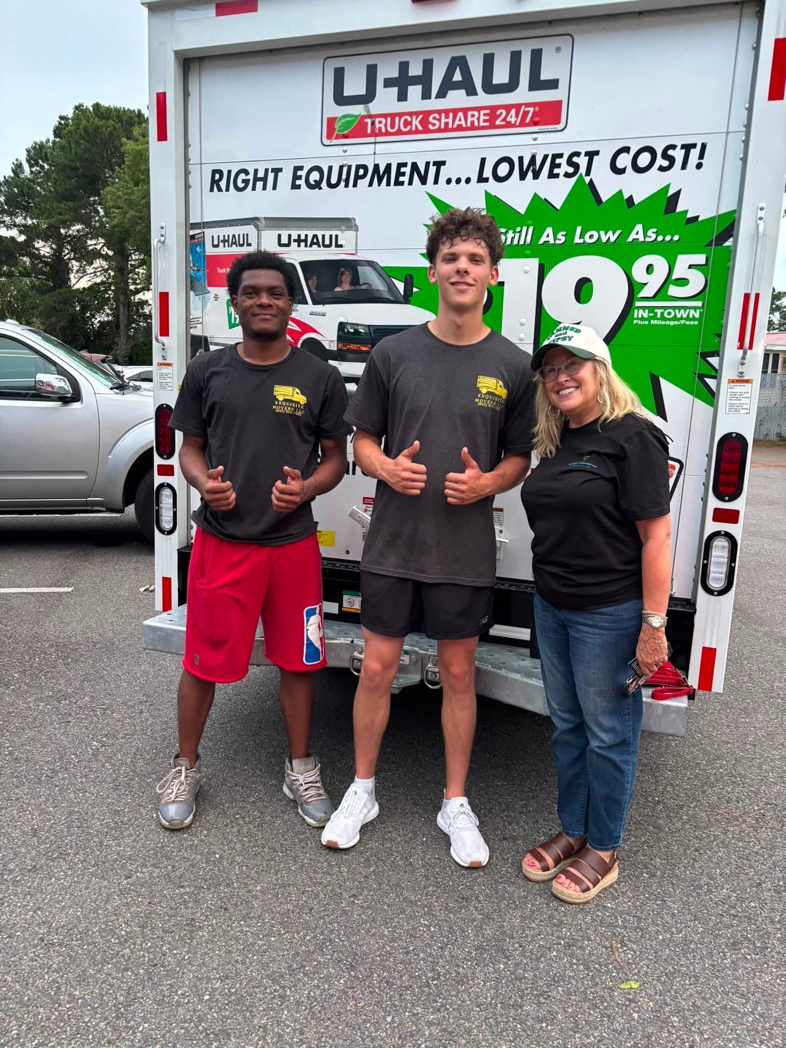 Three people posing in front of a U-Haul truck, smiling, with moving equipment and a rental sign behind them.