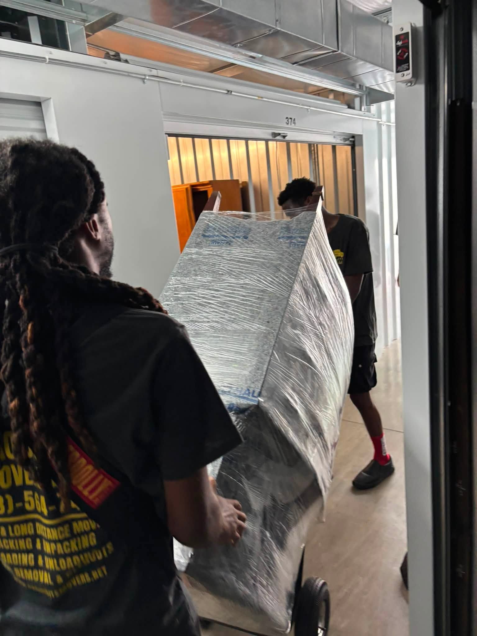 Two people carrying a large silver-wrapped object down a hallway with an orange-lit room ahead