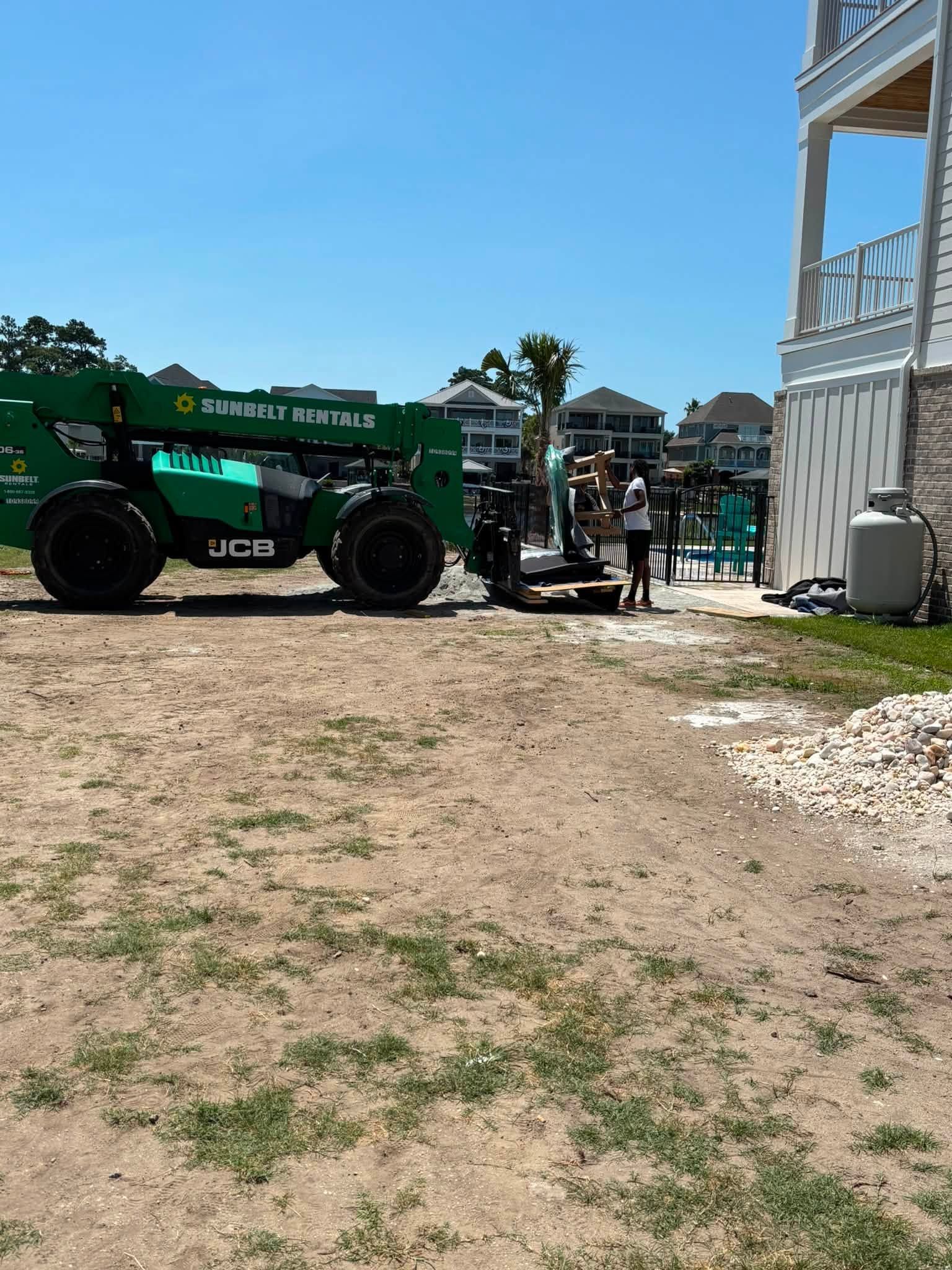 Green tractor by a fence near a sandy path, with people gathered by a waterfront dock under a blue sky