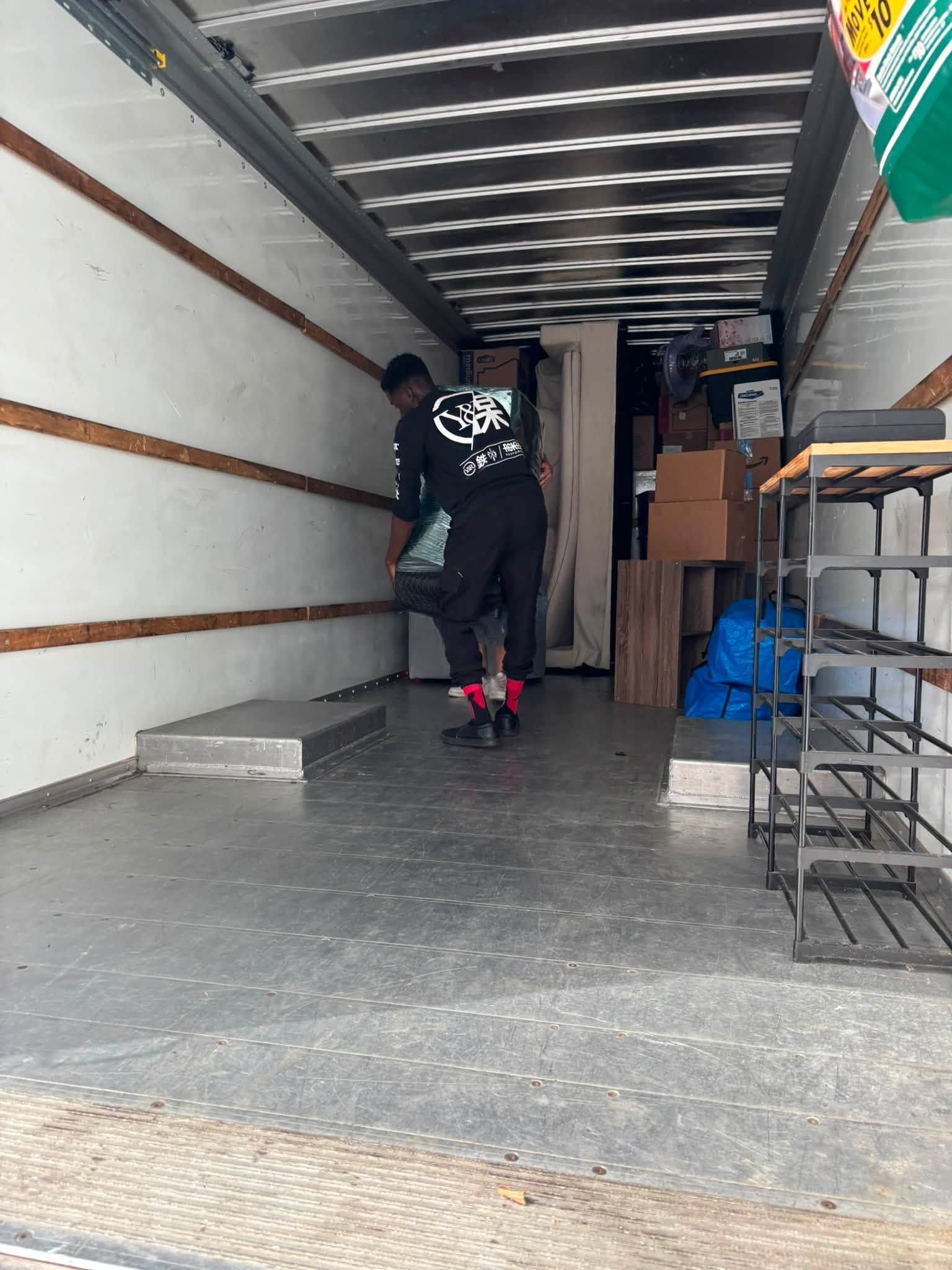 Person carrying boxes inside a moving truck, with a blue dolly and stacked cardboard boxes nearby.