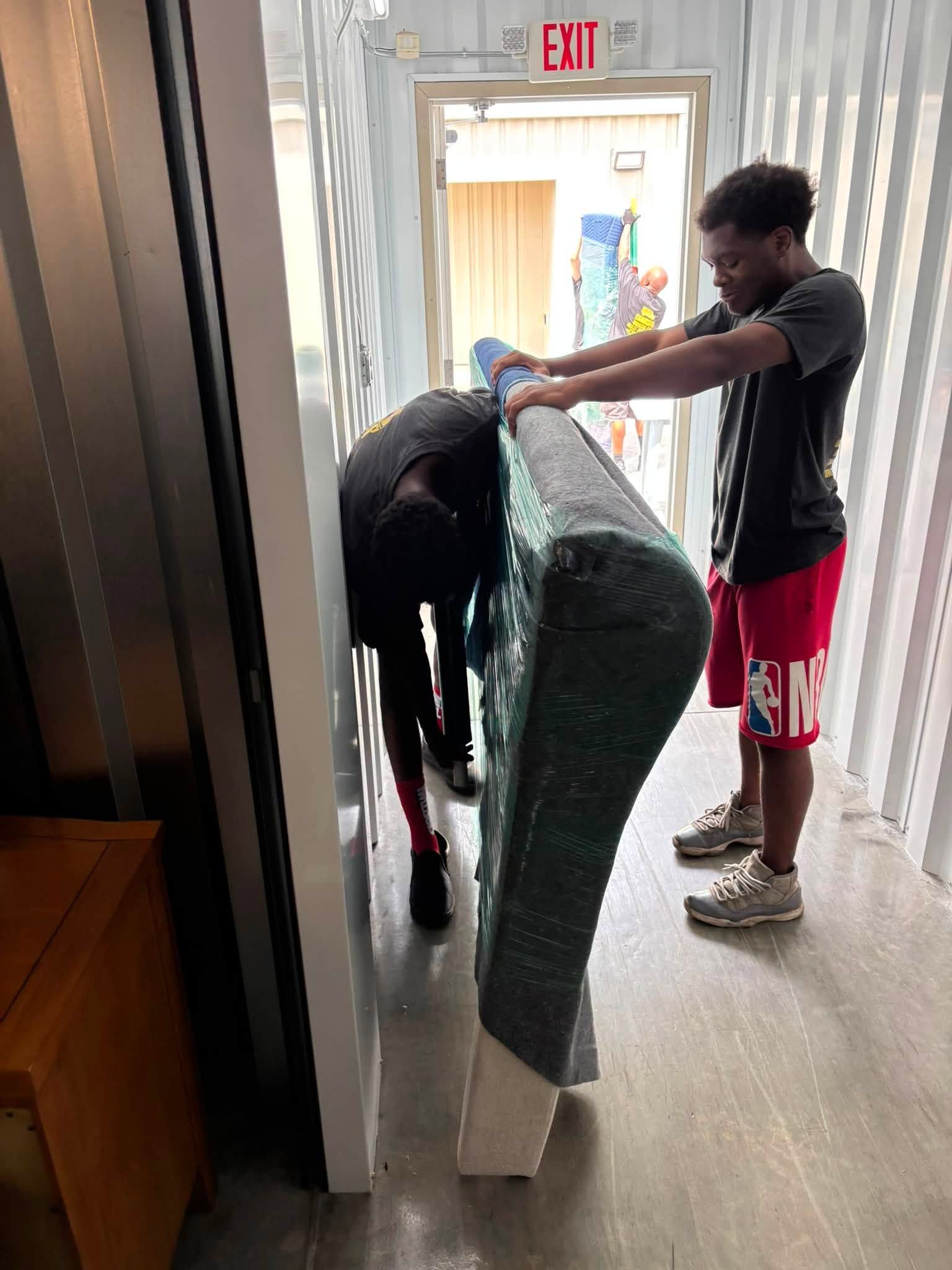 Two people carrying a green mattress through a doorway in a hallway