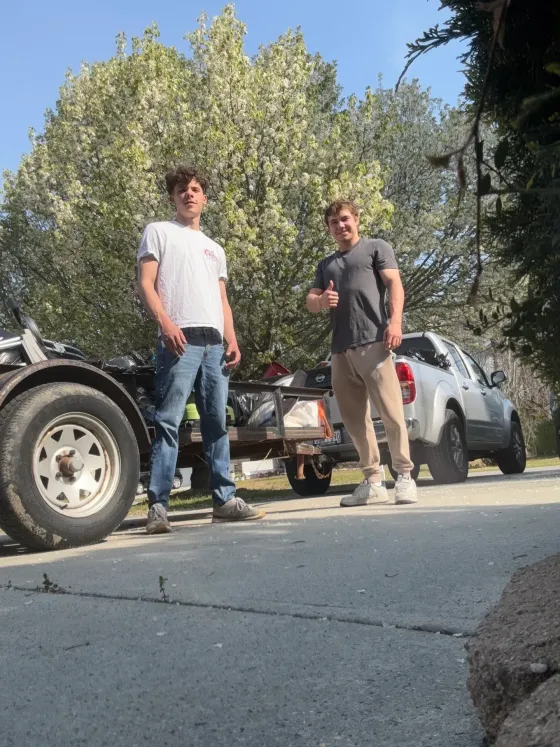 Two men standing by a car and trailer on a cracked road, with trees in the background.