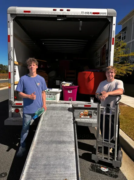Two people stand by a truck loading ramp with bins and a dolly in a sunny outdoor setting