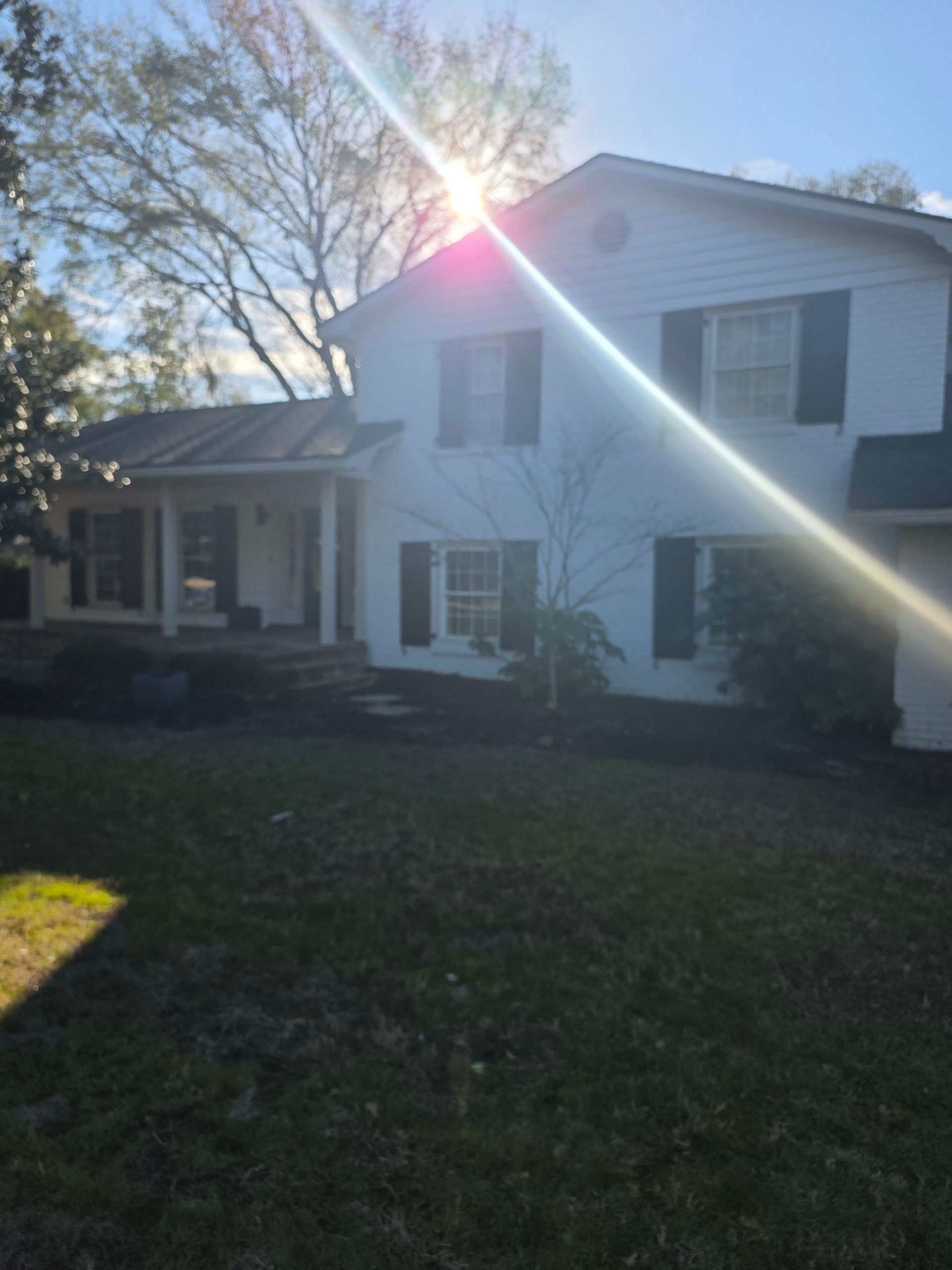 Two-story white house with black shutters and sun glare over the roof, front lawn in foreground