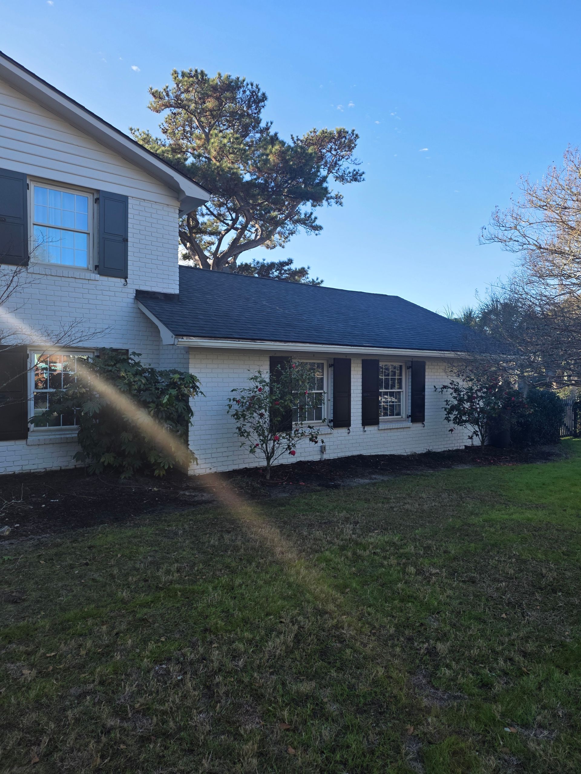 White two-story house with blue shutters and roof, sunlit yard, and trees in the background