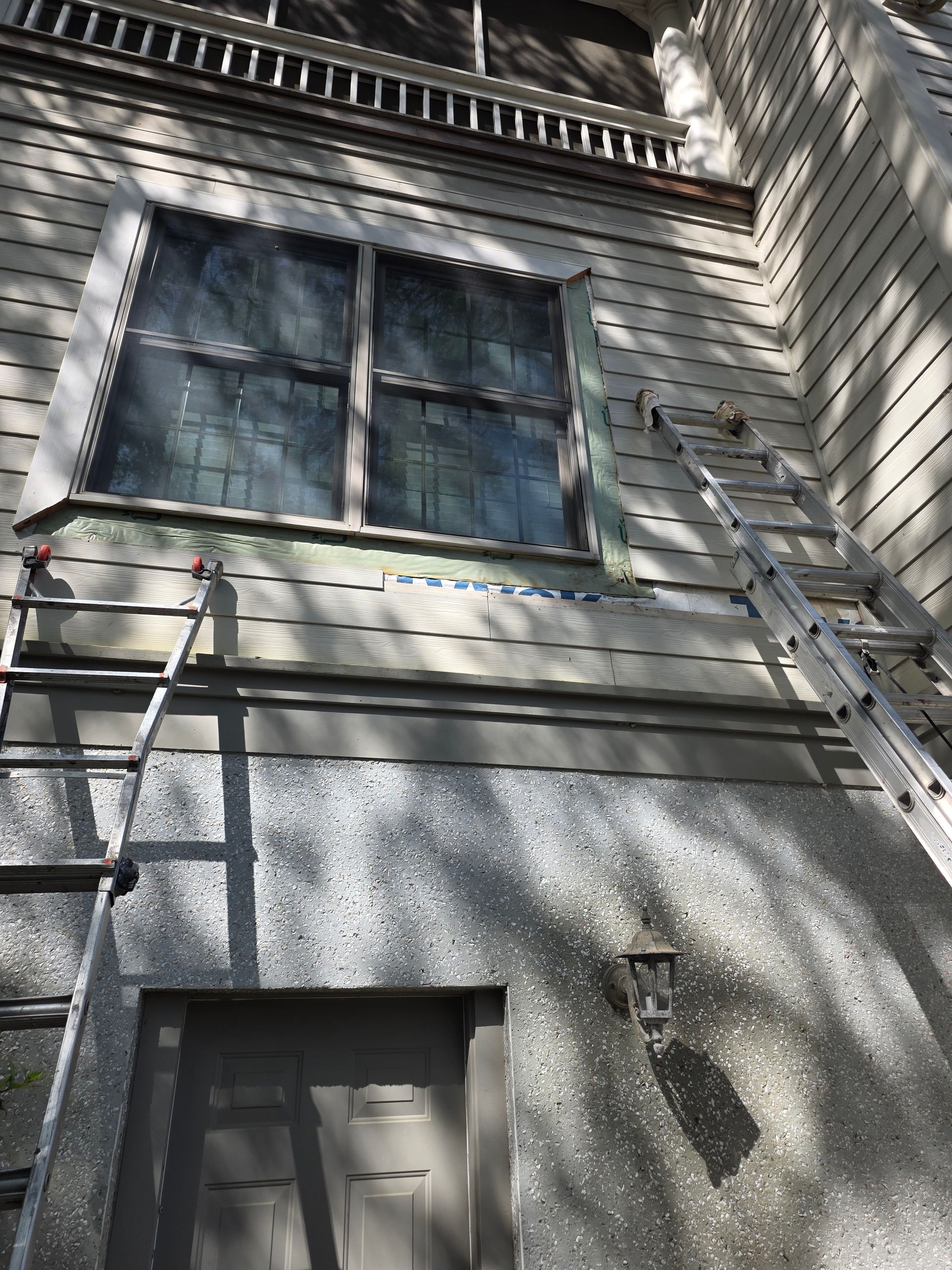 Ladders leaning against a gray house beneath a second-story window in sunlight
