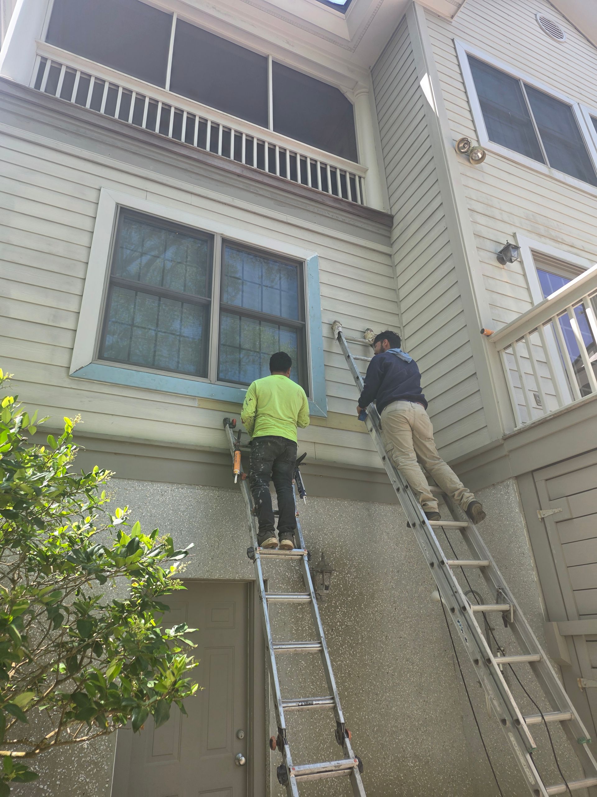 Two workers on ladders painting or repairing the exterior of a house under a window.