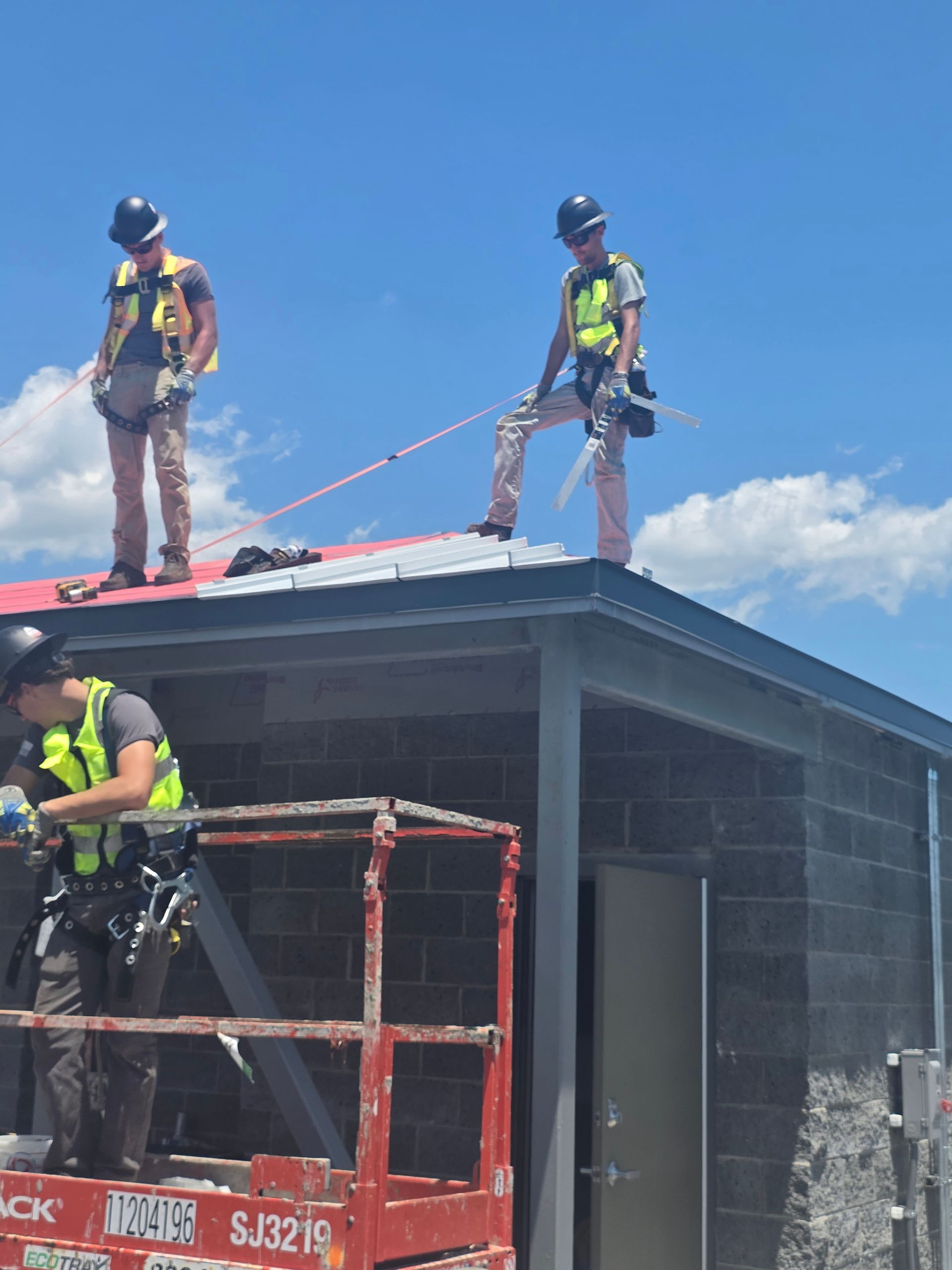 Workers on a rooftop using ropes above a red lift, with a cinder block building under a blue sky.