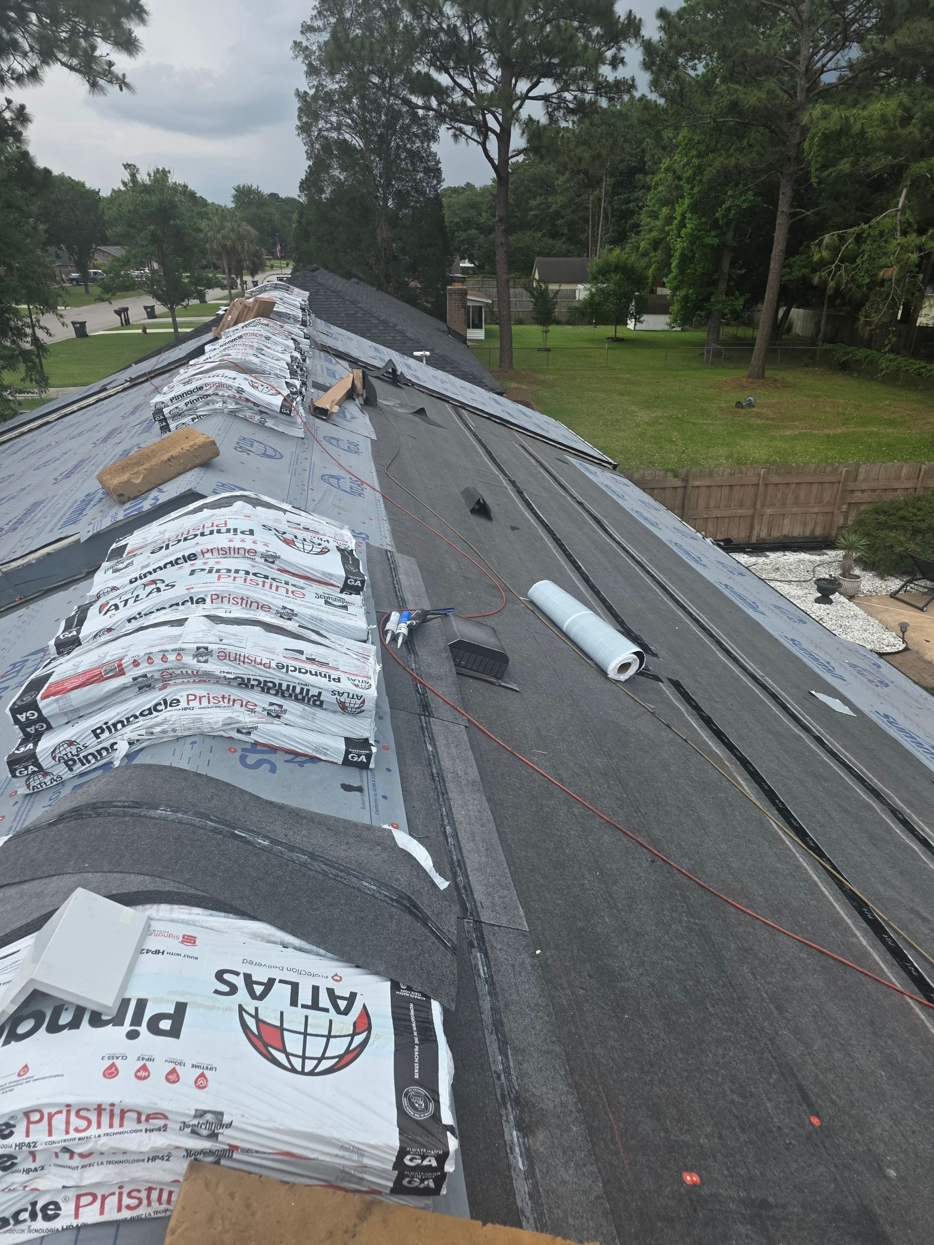 Damaged shingled roof with underlayment exposed in a wooded neighborhood after storm damage