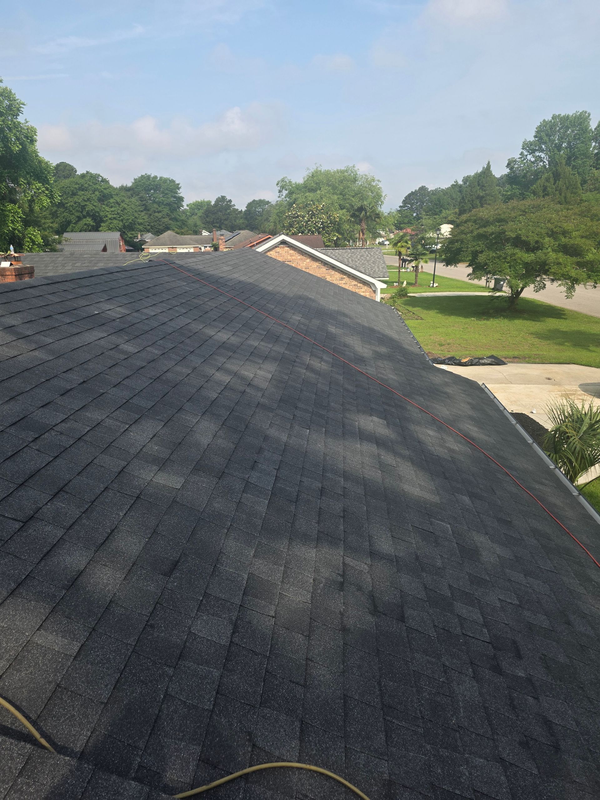 Dark shingled roof in foreground with trees and a suburban yard in the background