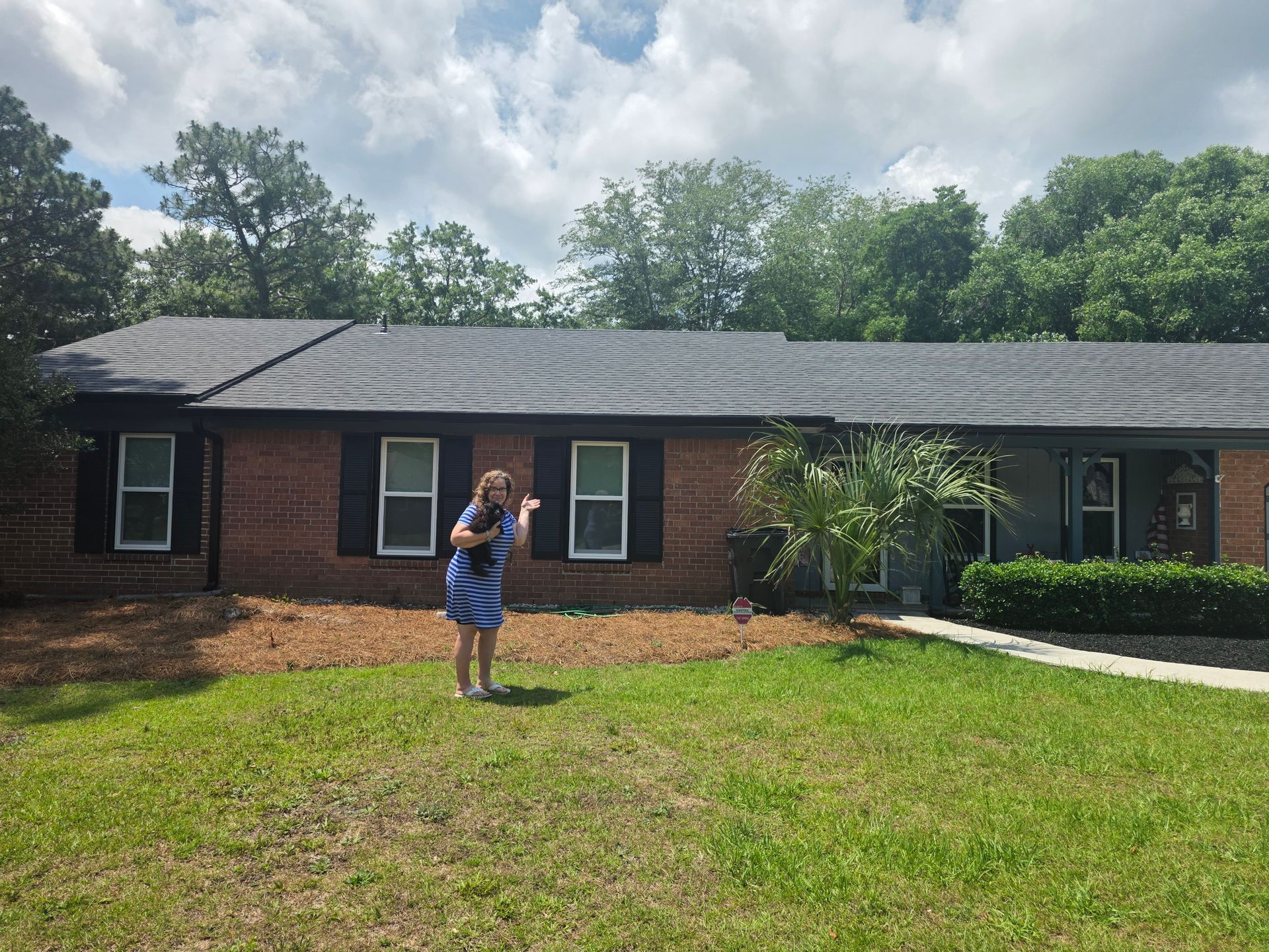 Person standing on lawn in front of a single-story brick house with trees and cloudy sky