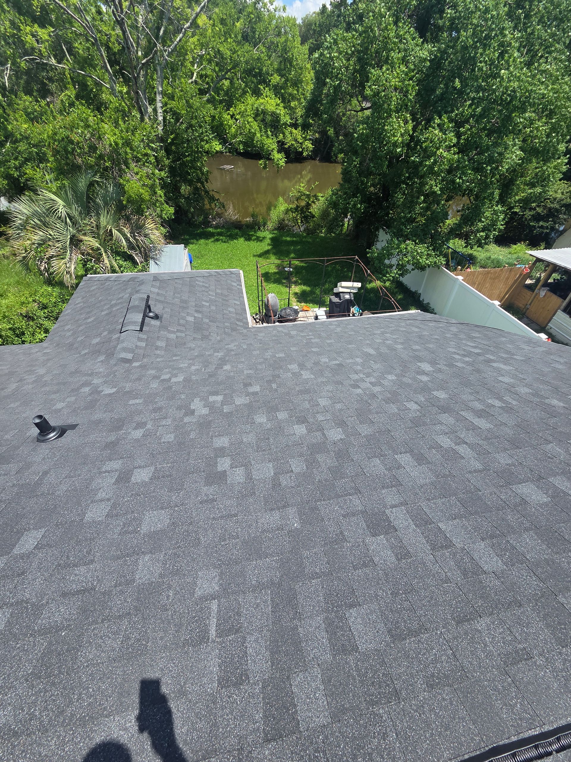 View from a gray shingled roof toward dense green trees and a small backyard patio below