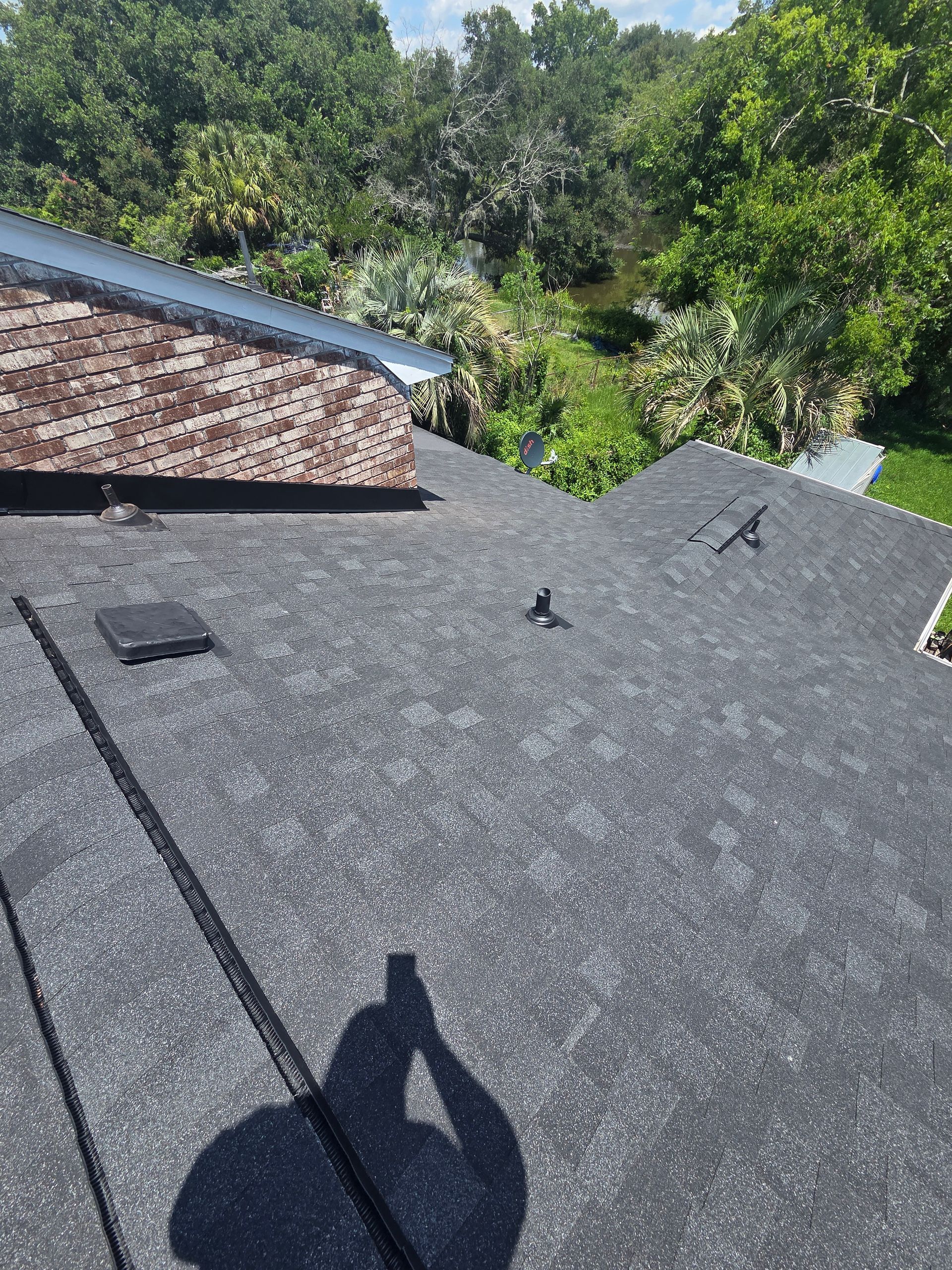 Rooftop covered in dark shingles with a vent and trees in the background.