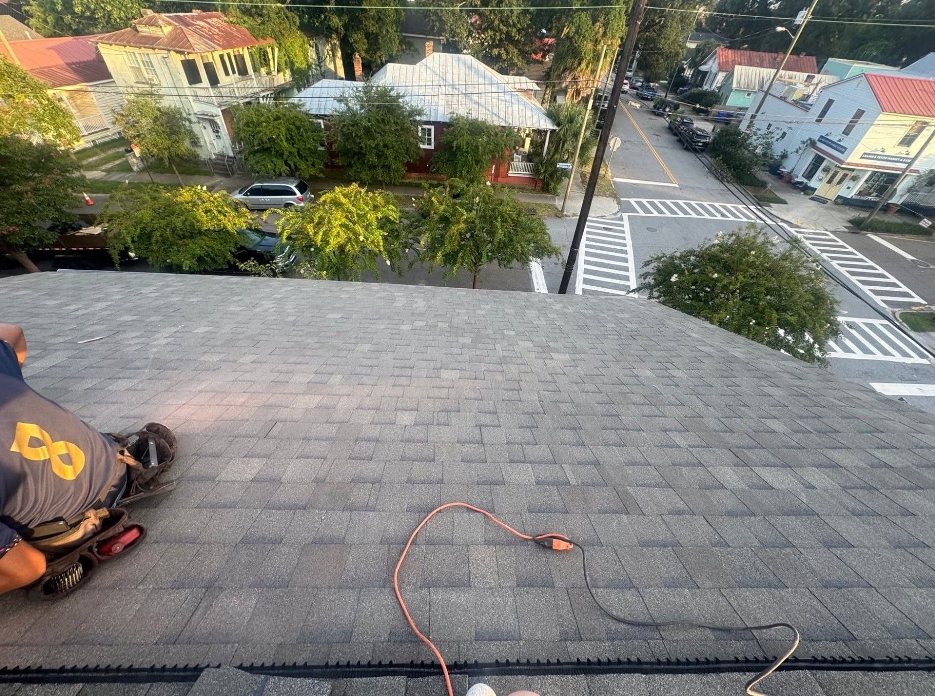 Roof edge overlooking a tree-lined street with parked cars and a crosswalk; extension cord on shingles