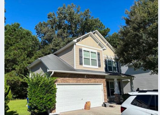 Suburban two-story beige house with white garage and trees under a bright blue sky