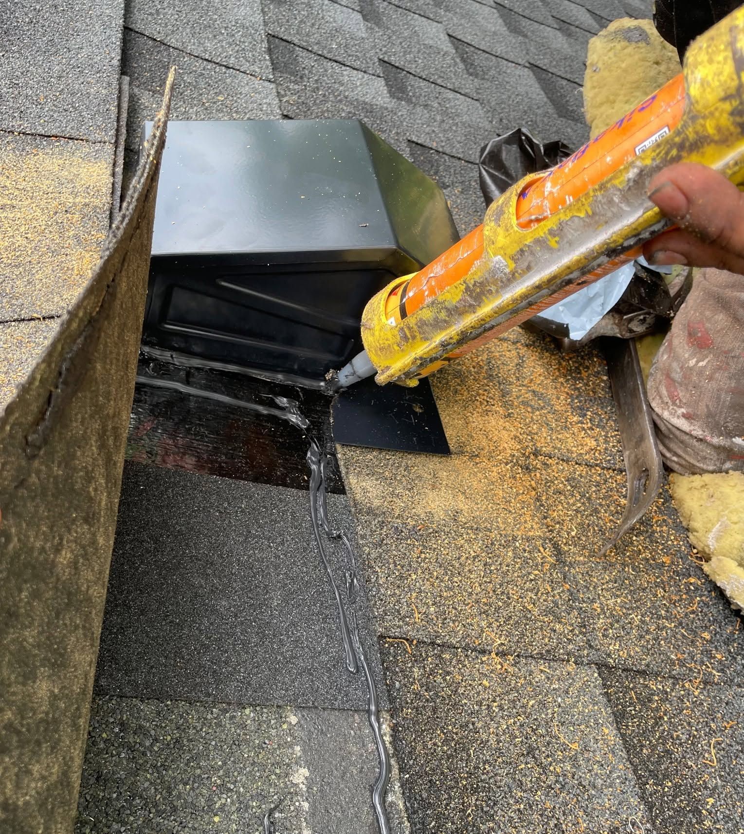 Worker applying yellow roofing sealant around a black roof vent on shingled roof