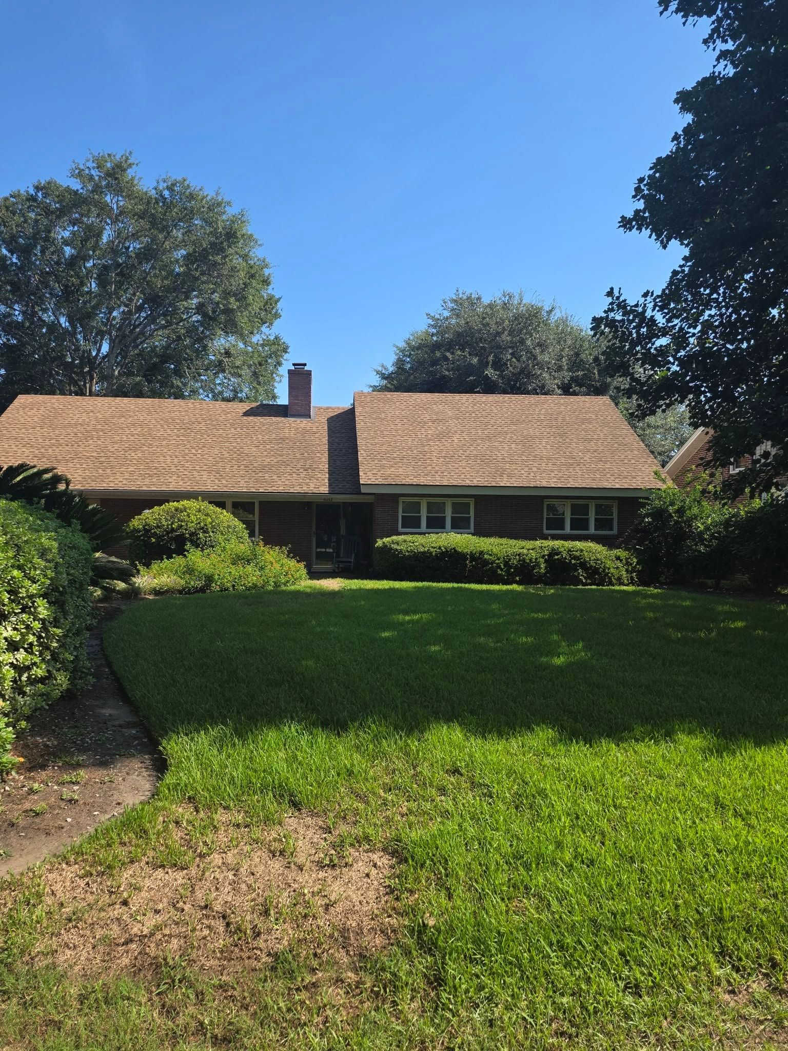 Suburban house with tan roof, green lawn, and trees under a clear blue sky