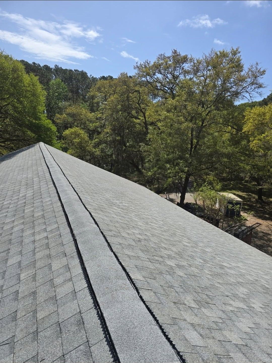 Gray shingled roof under a blue sky with trees and a yard in the background