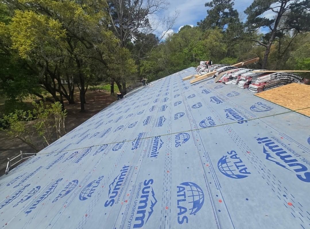 Roof covered with blue Atlas underlayment on a house, with trees and stacks of shingles nearby
