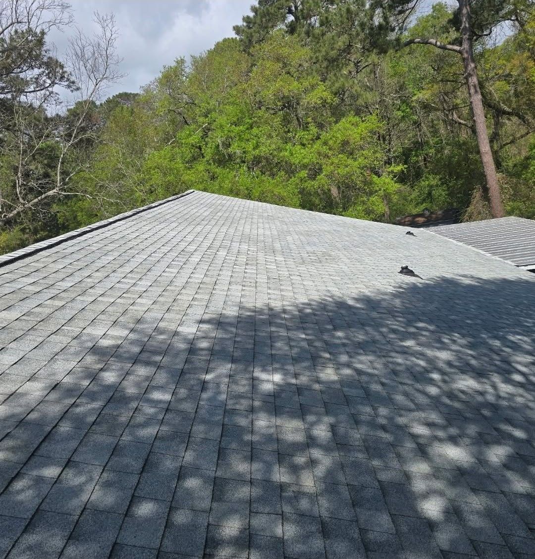 Gray shingled roof in a wooded area with trees and shadows under a partly cloudy sky