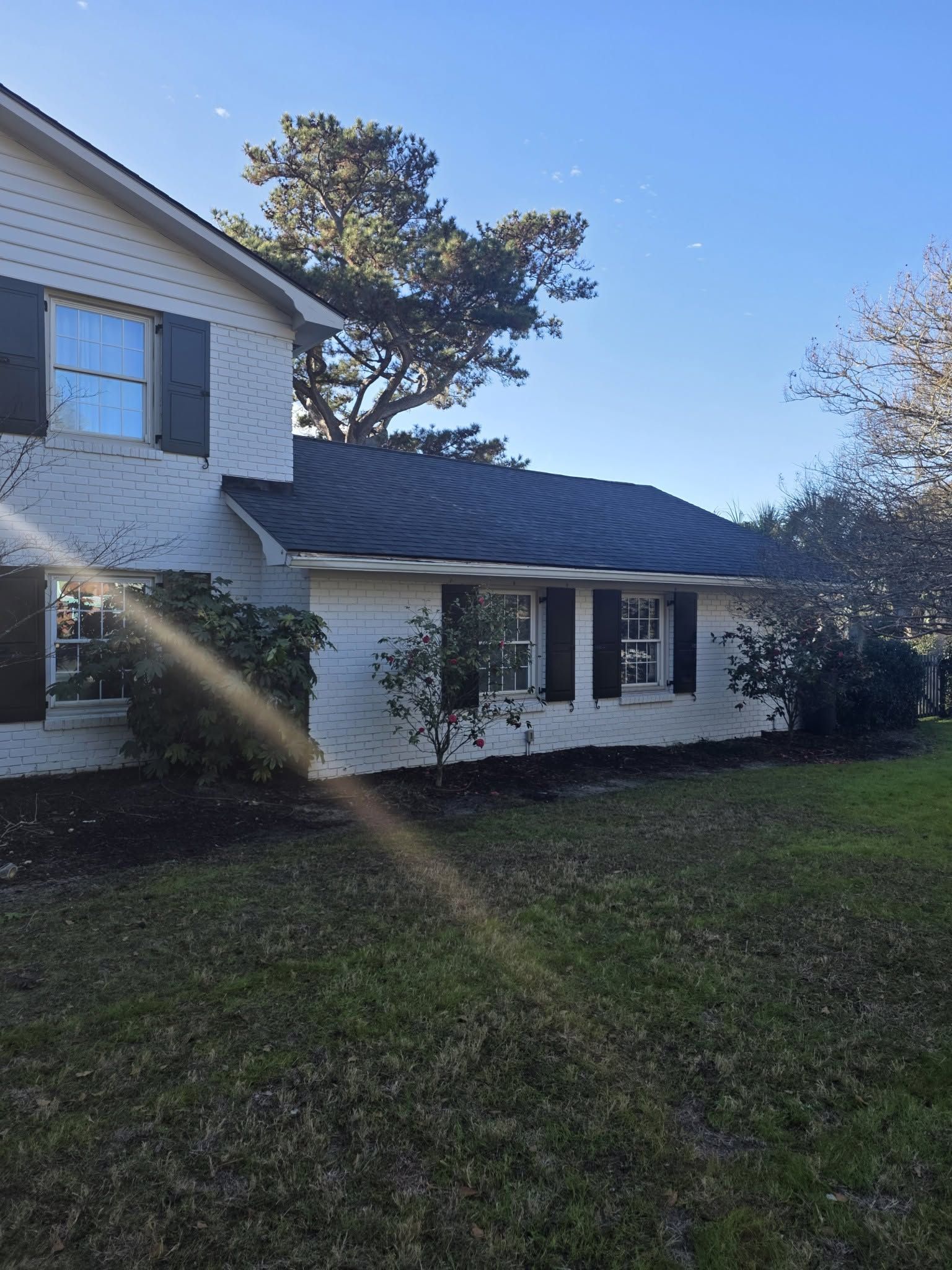 White two-story house with black shutters and a dark roof, seen from a grassy yard in bright sunlight